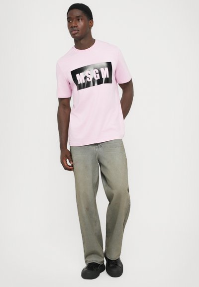 Mann iført en lys rosa MSGM-merke T-skjorte, løse grå jeans og svarte sko, som står foran en enkel hvit bakgrunn.