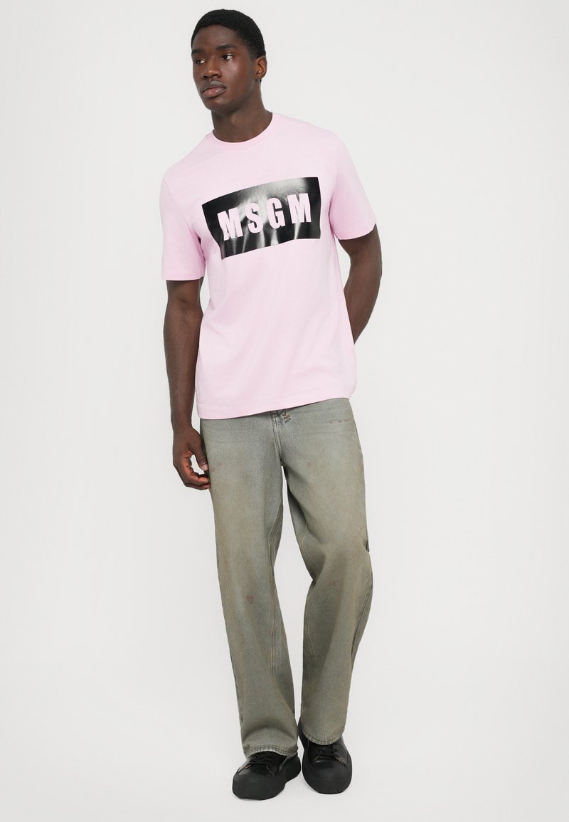 Man wearing a light pink MSGM branded T-shirt, loose gray jeans, and black shoes, standing against a plain white background.