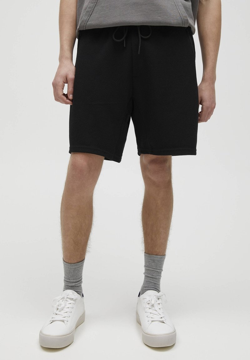 Shorts de sport noirs en tissu texturé, avec une taille élastique et des poches latérales, associés à des baskets blanches et des chaussettes grises.