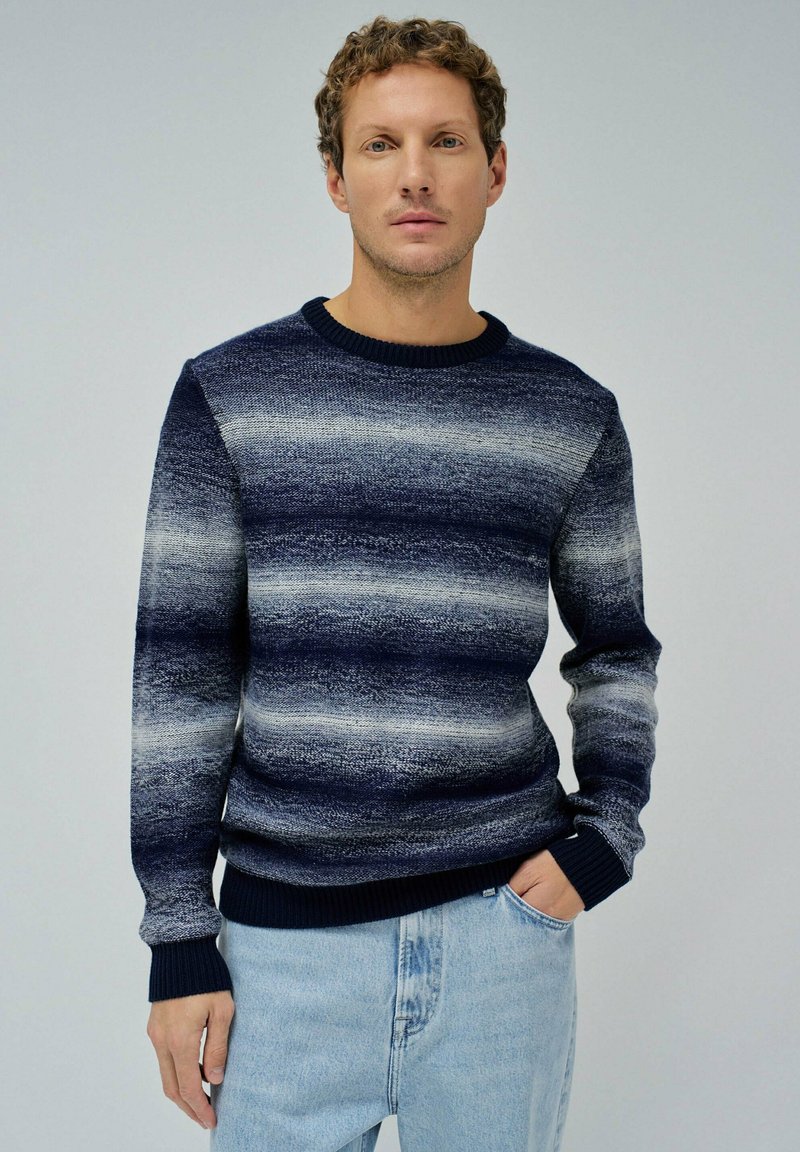 Suéter de punto a rayas en azul marino y gris con puños y cuello acanalados. La modelo lo lleva con jeans de un azul claro, con las manos en los bolsillos.