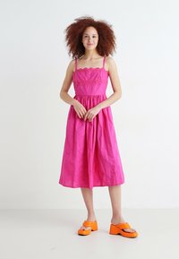 Rochie roz de lungime medie, cu un corset texturat, bretele subțiri și fustă plisată; purtată cu sandale cu platformă portocalii. Material neted, ușor.