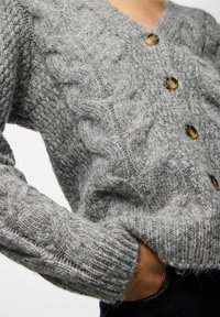 Pelēks adīts kardigans ar kabeli, ar V-veida kaklu, sešām brūnām pogām un ribotām piedurknēm. Maiga tekstūra ar mājīgu izskatu.