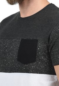 T-shirt med korte ærmer, der har en sort topsektion, en spottet sort og hvid krop og en sort lomme detalje på venstre side.