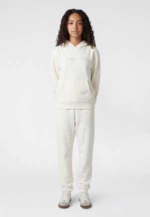 Giovane donna con capelli ricci che indossa una felpa Champion color avorio e dei pantaloni coordinati, in piedi con le mani dietro la schiena su uno sfondo semplice.