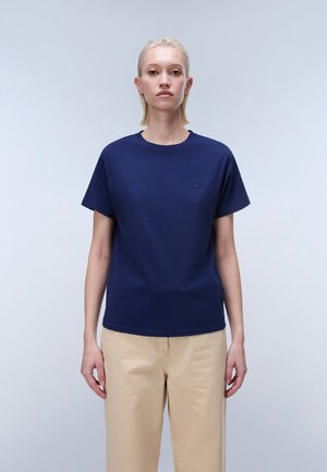 Frau mit kurzen blonden Haaren, die ein marineblaues T-Shirt mit kurzen Ärmeln und beige Hosen trägt, steht vor einem hellgrauen Hintergrund.