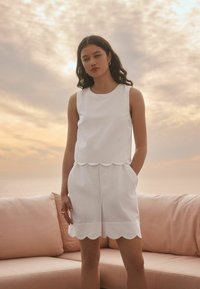 Haut sans manches blanc et shorts à ourlet festonné en tissu léger. Coupe décontractée avec poches latérales, présentés sur un fond pastel doux.