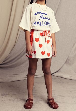 T-shirt crème avec texte bleu, associé à une jupe blanche à motifs de cœurs rouges et un cordon rouge. Des sandales marron complètent la tenue.