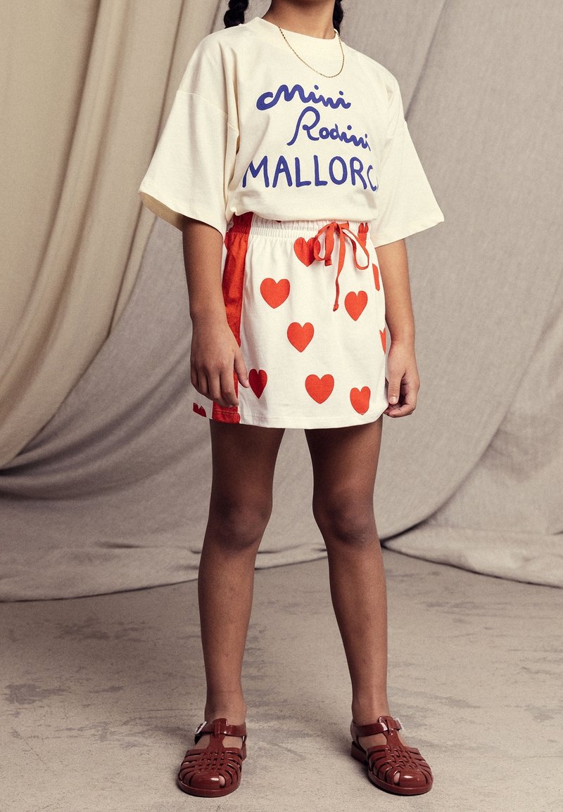 Crème t-shirt met blauwe tekst, gecombineerd met een witte rok met rode hartpatronen en een rode trekkoord. Bruine sandalen maken de outfit compleet.