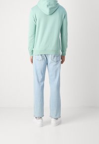Mátově zelená mikina s velkou kapucí, volný střih; kombinována se světle modrými džínami s rovnými nohavicemi a bílými teniskami. Jednoduchý design, bez viditelných vzorů.