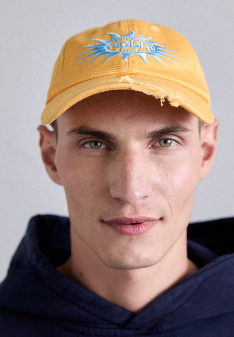 Gul baseball cap med et blåt grafisk logo og slidser på skyggen. Bruges med en marineblå hoodie, der har en blød tekstur og en afslappet pasform.