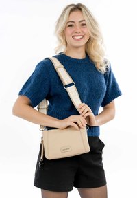 Beige crossbody bag with a textured leather finish, featuring a durable strap and a zippered closure, held by a person wearing a blue sweater.