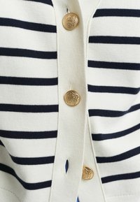Cardigan blanc avec des rayures navy, featuring des boutons dorés, une texture côtelée et un petit détail en forme de clé à l'avant.