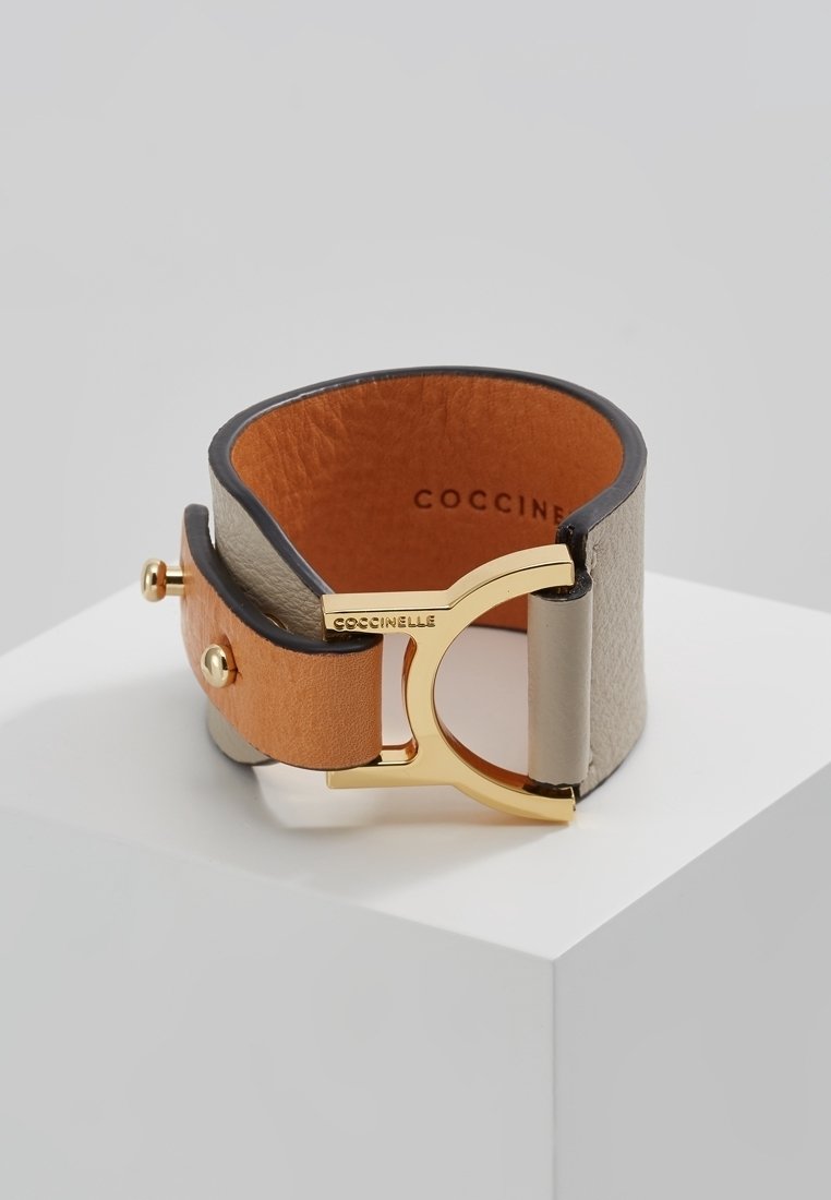 Large bracelet manchette en cuir bicolore avec fermoir en métal doré gravé "Coccinelle", présenté sur un piédestal blanc sur fond gris.