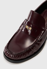 Bordeaux leren loafers met een gestikte moccasin neus, voorzien van een gouden embleem en een gladde, glanzende afwerking. Zwarte rubberen zool.