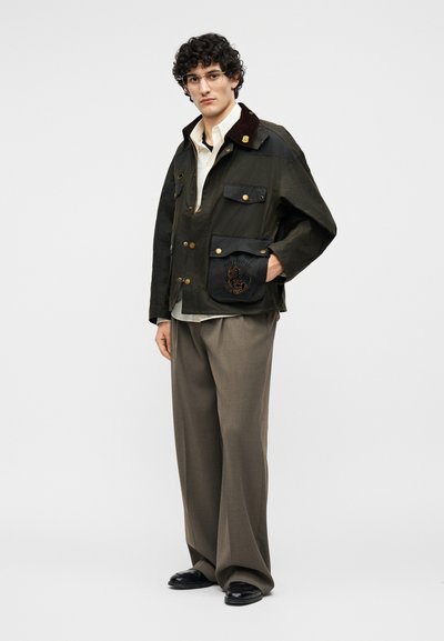 Άνδρας με σγουρά μαλλιά και γυαλιά, φορώντας σκούρο πράσινο σακάκι με κουμπιά, μπεζ πουκάμισο, φαρδιά καφέ παντελόνια και μαύρα παπούτσια, στέκεται με το ένα χέρι στην τσέπη.