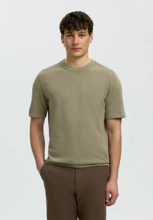 Jeune homme aux cheveux bouclés foncés portant un pull à manches courtes vert olive et un pantalon marron, debout avec une main dans la poche, devant un fond uni.