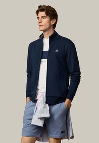 Jonge man die een marineblauwe jas met rits draagt, een wit met marineblauw gestreept shirt, lichtblauwe korte broek en een gestreept shirt om zijn middel geknoopt.