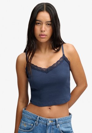 Femme aux longs cheveux foncés portant un haut sans manches bleu orné de dentelle et un jean bleu ample, les mains derrière le dos.