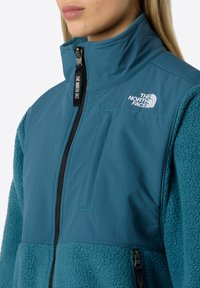 Tyrkysová fleecová bunda se zapínáním na zip, hladkými horními panely a texturovanými dolními částmi. Obsahuje logo na hrudi a kapsu na zip po straně.