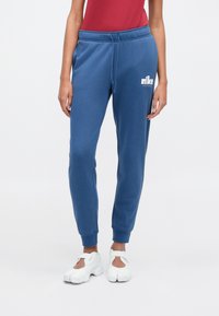 Modré tepláky Nike z mäkkej látky. Majú elastický pás so šnúrkou, manžety na nohaviciach a biely logo na ľavom stehne.