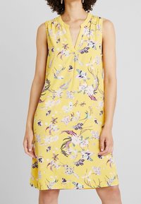 Ärmelloses floral bedrucktes Kleid in strahlendem Gelb, mit V-Ausschnitt, gefältelten Akzenten und bunten Blumenmustern überall.