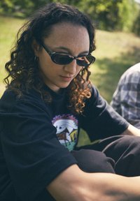 Camiseta preta com um design gráfico colorido, óculos de sol curvos e cabelo encaracolado; o cenário inclui relva verde e um fundo desfocado.