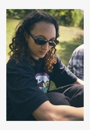 Must t-särk, millel on värvikas graafiline kujundus, kõverad päikeseprillid ja lokkis juuksed; taustal roheline muru ja hägune taust.
