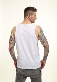 Camiseta sin mangas de algodón blanco con cuello redondo. Presenta tatuajes coloridos en el brazo izquierdo y jeans grises.
