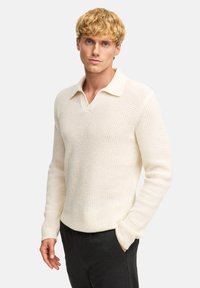 Maglione lavorato a maglia di colore crema con scollo a V e colletto, caratterizzato da un motivo testurizzato, maniche lunghe e una vestibilità ampia. Indossato sopra pantaloni scuri.