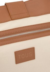 Brązowy pasek z imitacji skóry na kremowej, teksturowanej torbie z tkaniny, z widocznym srebrnym zamkiem błyskawicznym i prostokątną, jasnobrązową metką z tłoczonym napisem "TOUS".