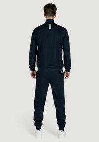 Tuta da jogging navy realizzata in tessuto liscio. Presenta un colletto alto, pantaloni affusolati e un dettaglio a strisce sul retro. Sneakers bianche completano il look.