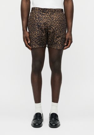 Man draagt bruin-zwart leopardprint korte broek, witte sokken, zwarte loafers en een wit overhemd, staand tegen een effen achtergrond.