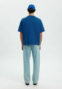Blå kortærmet T-shirt med en afslappet pasform, parret med lyseblå denimbukser, set bagfra mod en ensfarvet hvid baggrund.