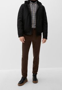 Zwarte zip-up jas, geblokt shirt in blauw en wit, bruine broek en zwarte sneakers met een bruine zool, weergegeven tegen een effen achtergrond.