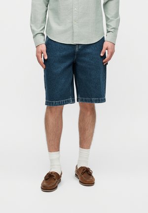 Mees, kes kannab nööbitud halli särki, siniseid teksapükse, valgeid sokke ja pruune paadijalatsid, seisab ühevärvilise tausta ees.