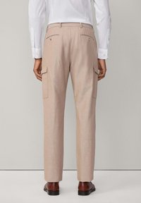 Beige linnebyxor med två sidofickor, bakfickor med knappdetaljer och slim fit design, kombinerade med bruna skor.