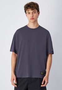 Przeciętny, dużych rozmiarów, szary t-shirt wykonany z bawełny, z krótkimi rękawami, okrągłym dekoltem oraz boczną metką z żółtym napisem i guzikiem.