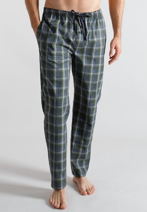 Man wearing green and blue plaid pajama pants with black drawstring, barefoot, standing against plain light background.
