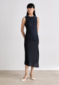 Navy blue sleeveless dress with textured fabric, asymmetrical design, side slit, and a fitted silhouette, paired with white pointed heels.