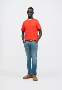 Punainen lyhythihainen t-paita pyöreällä pääntiellä, siniset farkut ja ruskeat mokkanahkakengät. Paidassa on pieni logo rinnassa.