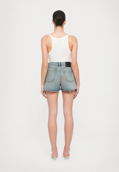 Jeanshorts med rå kant, lys blå vask, middels høy liv og forsterket søm. Brukt med en hvit tanktop og nøytrale hæler.