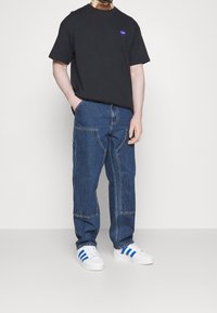 Camiseta de algodón negra con un logo de corazón morado, combinada con pantalones de mezclilla azul con bolsillos parches. Zapatillas blancas con rayas azules.