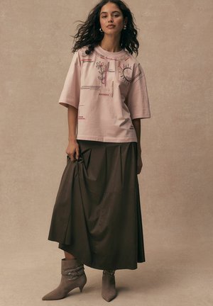 Woman with curly hair wears a pale pink embroidered shirt, dark brown midi skirt, and taupe ankle boots with eyelet detailing.
