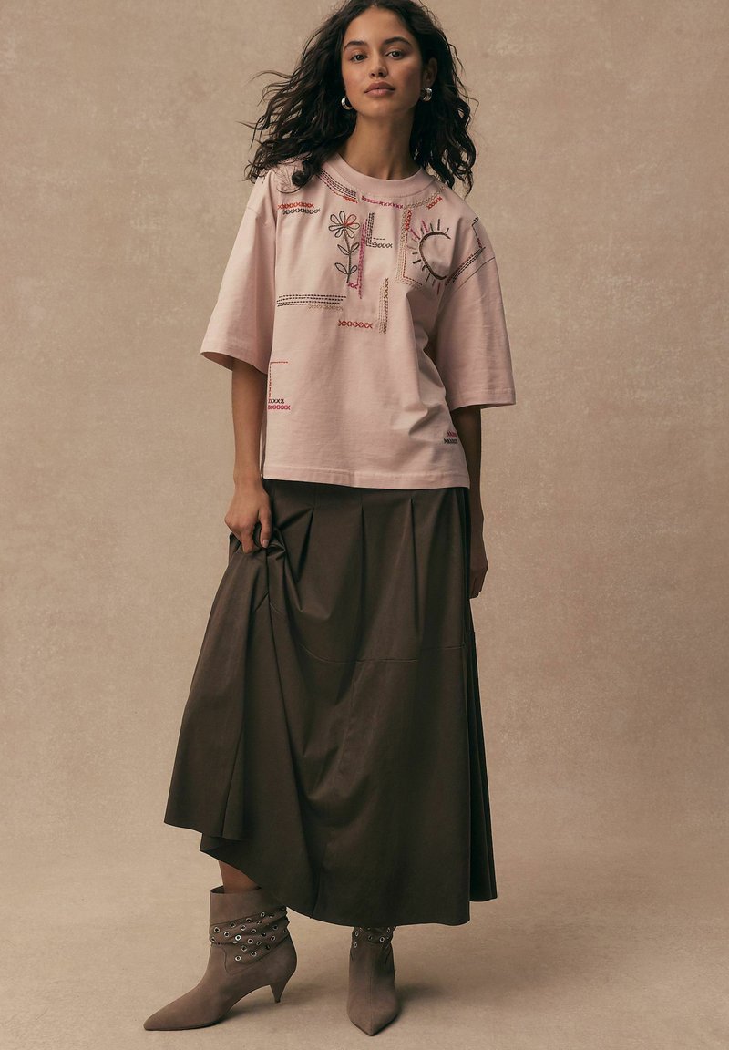 Kobieta z kręconymi włosami ubrana w bladoróżową haftowaną koszulę, ciemnobrązową spódnicę midi i taupe botki z ozdobnymi otworami.