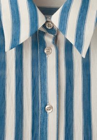 Gros plan d'une chemise à rayures verticales bleues et blanches avec trois boutons blancs visibles et un col pointu.