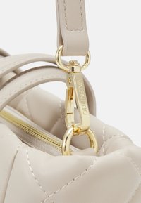 Beige quiltad handväska med en guldfärgad lås med en ögla. Texturerat läder och dragkedjedetaljer synliga på toppen.