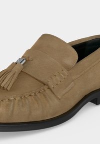 Ανδρικά μοκασίνια από καστανό σουέντ με λεπτομέρεια φουντίτσας, μαύρη σόλα και ορατές ραφές στην περιοχή των δαχτύλων.