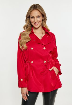 Frau mit langen welligen blonden Haaren, die eine knallrote zweireihige Jacke mit goldenen Knöpfen und schwarze Hose trägt und lächelt, mit einer Hand in der Tasche.
