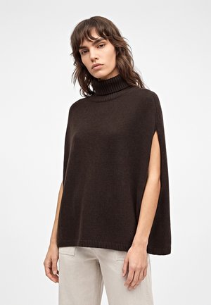 Жена, облечена в тъмнокафяв плетен пончо с поло яка и разрези за ръцете и светлобежови панталони на бял фон.