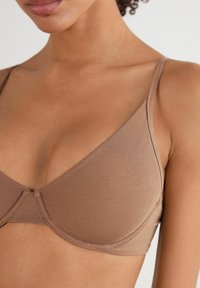Hnědá podprsenka s bezešvým designem, hladkým bavlněným materiálem a tenkými nastavitelnými ramínky, s jemně zakřiveným tvarem a žádným viditelným kováním.